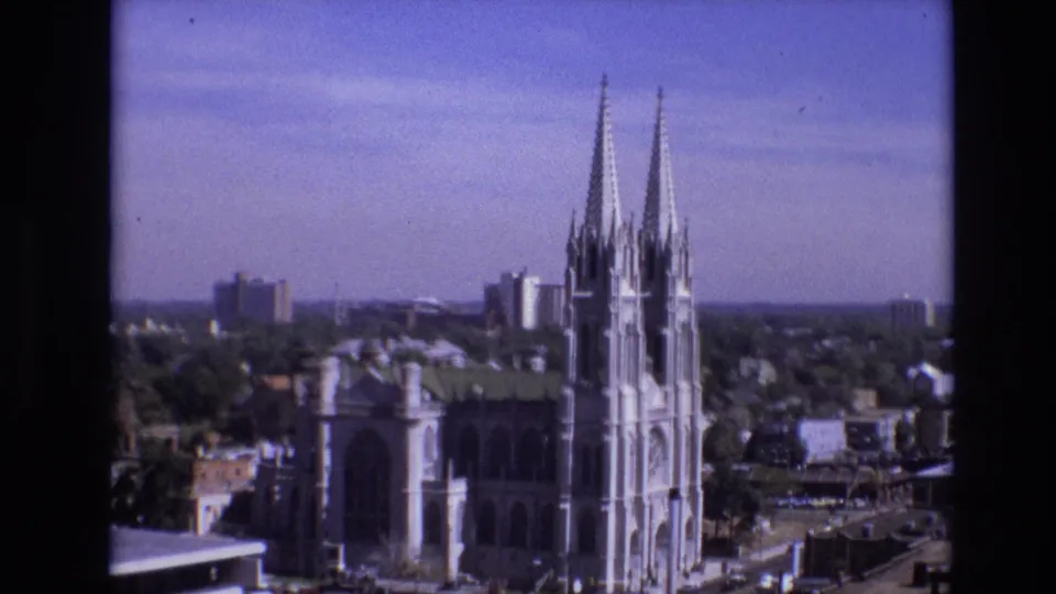 Wide 1974 Denver home-movie view showing the cathedral's twin spires and surrounding urban rooftops, with taller buildings in the distance.