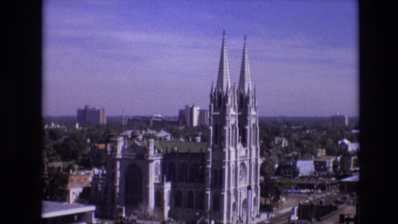 1974 Denver city view with a Gothic church, twin spires, surrounding downtown buildings, trees, and blue sky on 8mm film.