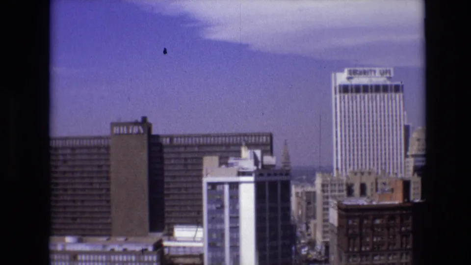 1974 Denver skyline view from a high vantage point, showing a Hilton hotel, the Security Life tower, nearby office buildings, and blue sky within an 8mm frame.