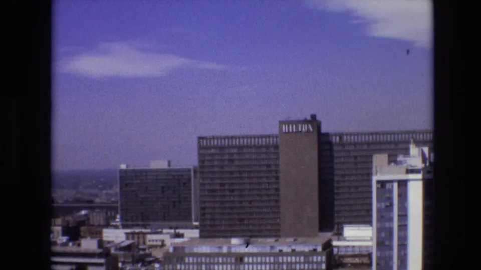 1974 Denver skyline frame with a large Hilton hotel block, a dark office building, a white tower at the right edge, rooftops in the foreground, and a broad blue sky.