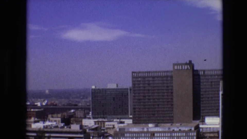 Final 8mm frame of the Denver downtown panorama, with the Hilton-branded tower at right, lower commercial buildings, distant cityscape, and scattered clouds overhead.