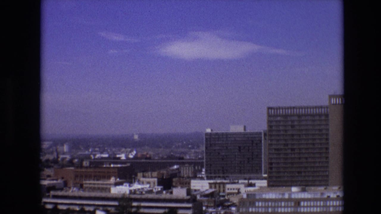 Elevated 8mm view across downtown Denver, showing mid-century towers, lower rooftops, and a hazy horizon beneath scattered clouds.
