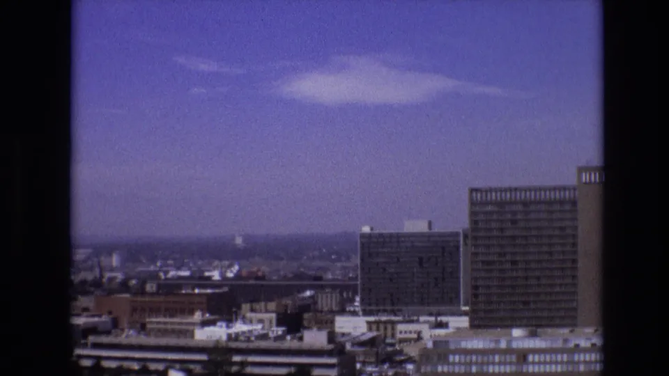 Elevated 8mm view across downtown Denver, showing mid-century towers, lower rooftops, and a hazy horizon beneath scattered clouds.