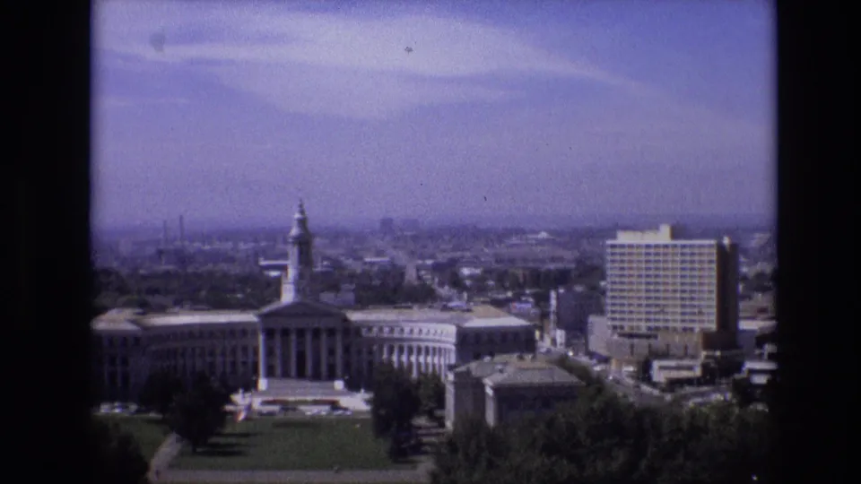 Denver skyline frame from 1974 showing a classical-style civic building, tree-lined foreground, roads, and a tall rectangular tower under a pale blue sky.