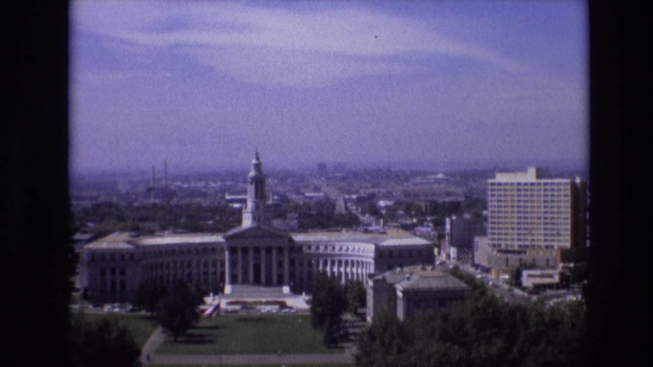 High-angle view of a classical civic building with a tower, columns, lawn, trees, and nearby downtown buildings.
