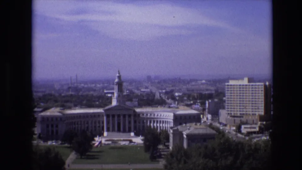 1974 Denver home-movie frame showing a high-angle view of a large classical-style civic building with a central tower, front columns, lawns, trees, and surrounding urban buildings.