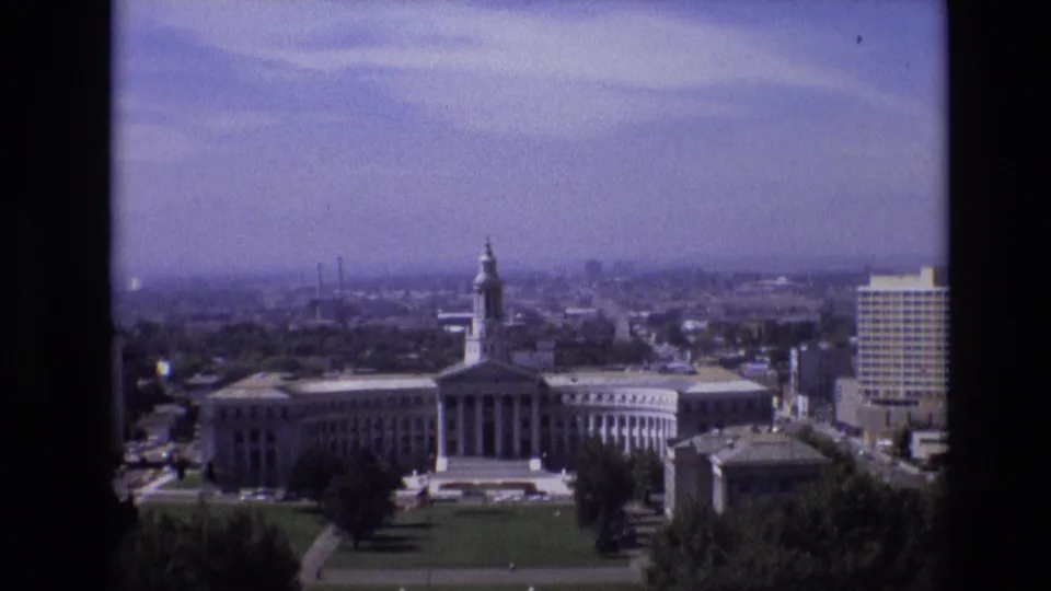1974 Denver frame with a broad elevated view of a towered classical civic building, formal lawn, scattered trees, distant city blocks, and a tall building at the right edge.