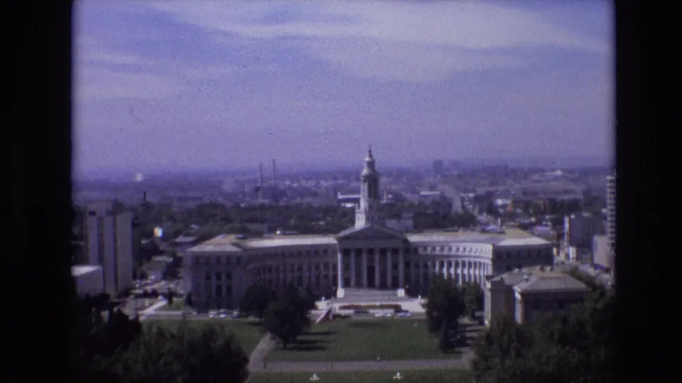 1974 Denver home-movie frame showing the camera shifted across the classical civic building, with its tower, long facade, green grounds, trees, and hazy urban skyline visible beyond.