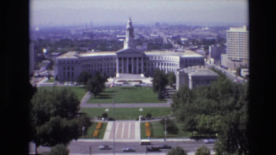 1974 Denver frame showing a closer elevated view of the classical civic building and grounds, with symmetrical walkways, green lawn, trees, a road with cars, and downtown buildings in haze.