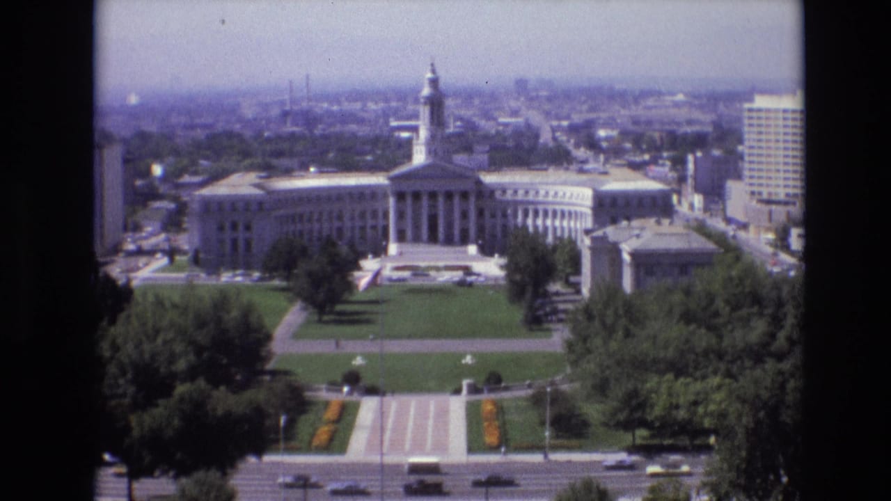 1974 Denver elevated view of a large white government building with classical columns and a central tower, bordered by green civic lawns, paths, trees, traffic, and downtown buildings.