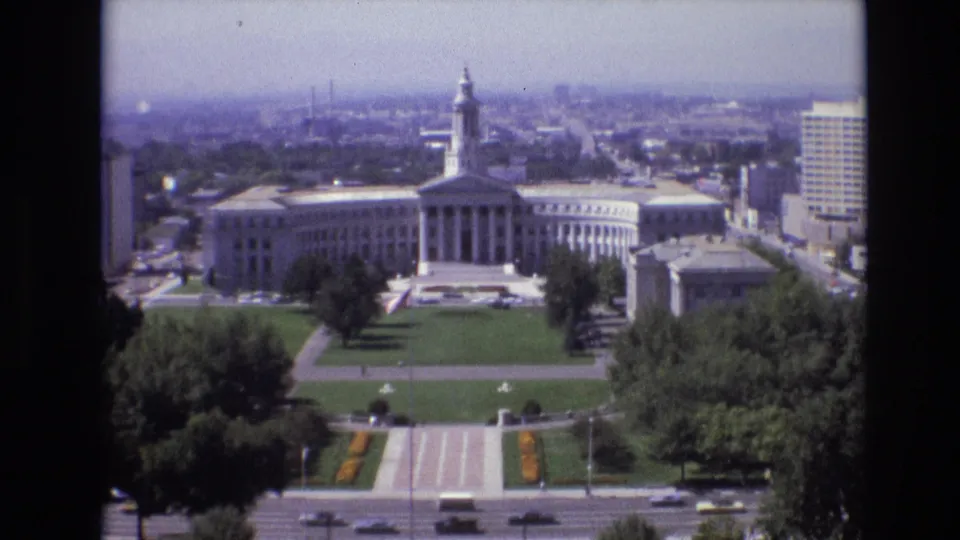 1974 Denver elevated view of a large white government building with classical columns and a central tower, bordered by green civic lawns, paths, trees, traffic, and downtown buildings.
