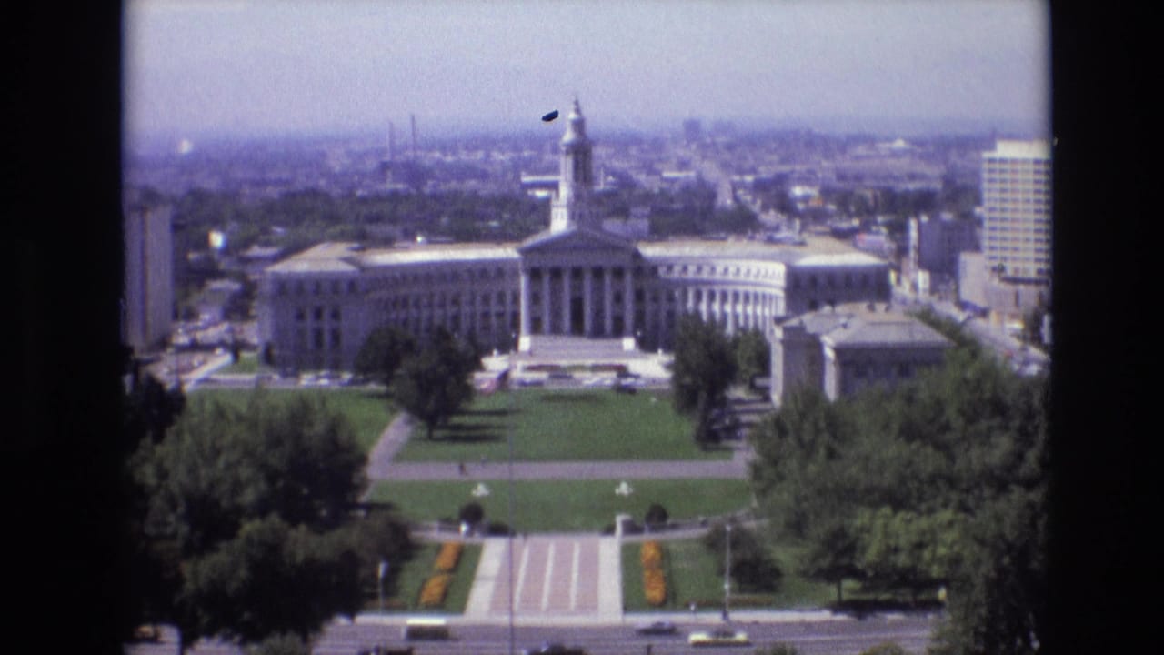 1974 Denver view looking across civic lawns and a street toward a white government building with columns and a tower, framed by trees, office buildings, and a hazy skyline.