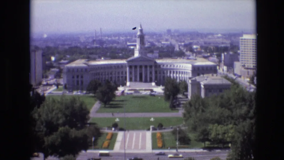 1974 Denver view looking across civic lawns and a street toward a white government building with columns and a tower, framed by trees, office buildings, and a hazy skyline.