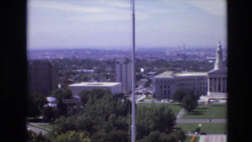 1974 Denver pan showing downtown buildings, a vertical pole in the foreground, civic lawns, trees, street traffic, and the partial edge of the white government building.