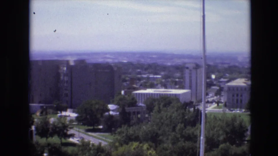 1974 Denver elevated city view with office blocks, civic lawns, trees, traffic, a foreground pole, and part of the white government building near the right edge.