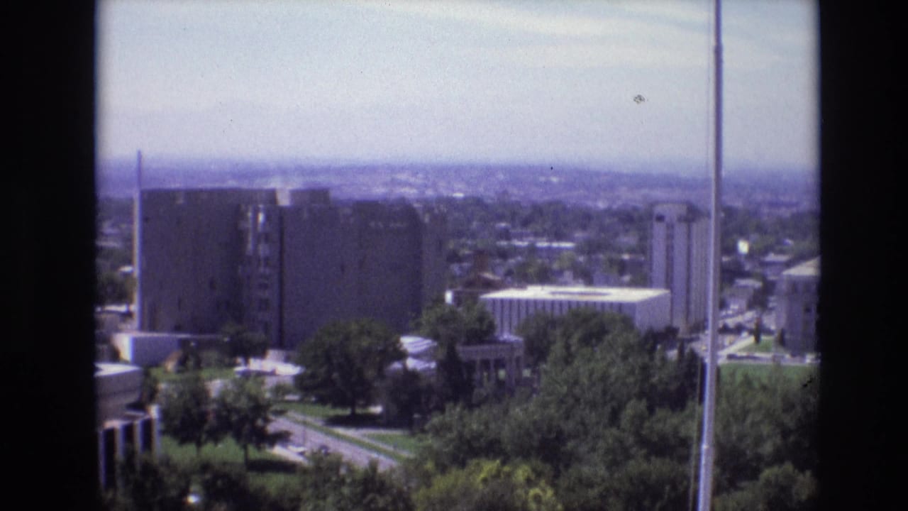 1974 Denver home-movie frame showing an elevated city view with trees, modernist buildings, hazy horizon, and dark balcony edges.