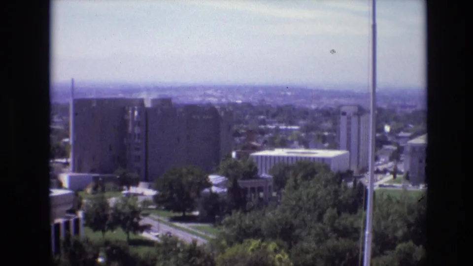1974 Denver home-movie frame showing an elevated city view with trees, modernist buildings, hazy horizon, and dark balcony edges.