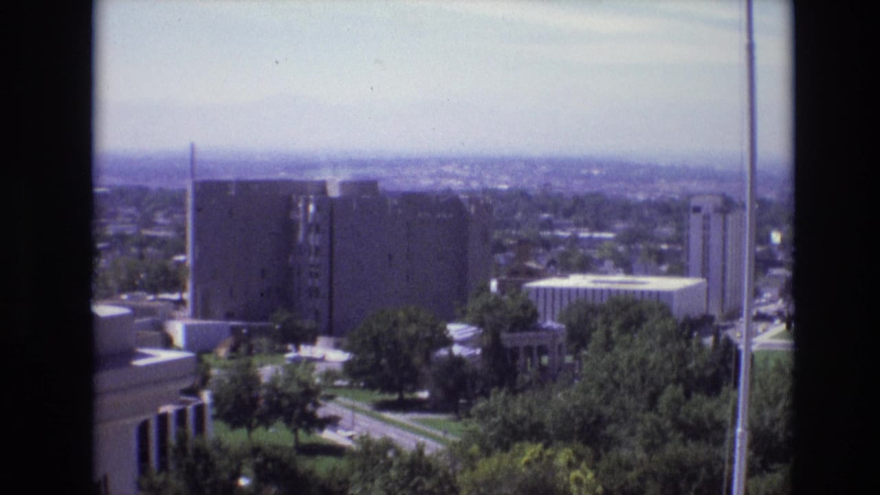 1974 Denver skyline view from an elevated balcony, with mid-century buildings, trees, a bright sky, and dark frame edges from the filming position.