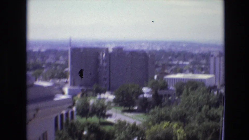 Soft 8mm frame of a 1974 Denver city overlook, with downtown structures, trees, balcony architecture at the edge, and visible film specks.