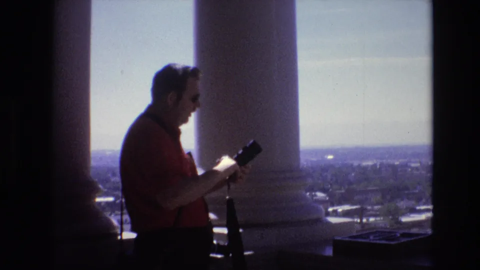 1974 Denver balcony scene with an adult photographer in a red shirt and sunglasses holding a camera beside white columns above the city view.