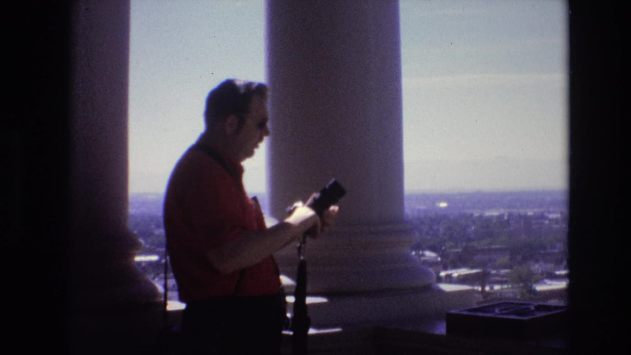 Man in a red shirt looks down at a camera between large white columns with a city view behind him.