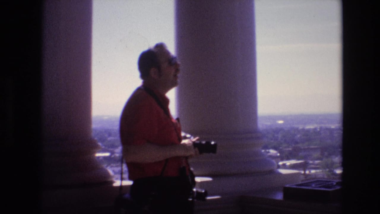 Man in sunglasses holds a camera while standing beside tall white columns above a distant cityscape.