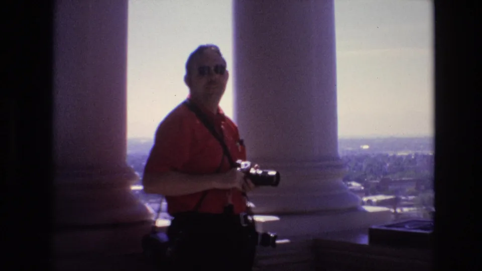 The man in a red shirt and sunglasses turns toward the 8mm camera with his still camera in hand, framed by white columns and the Denver skyline in 1974.