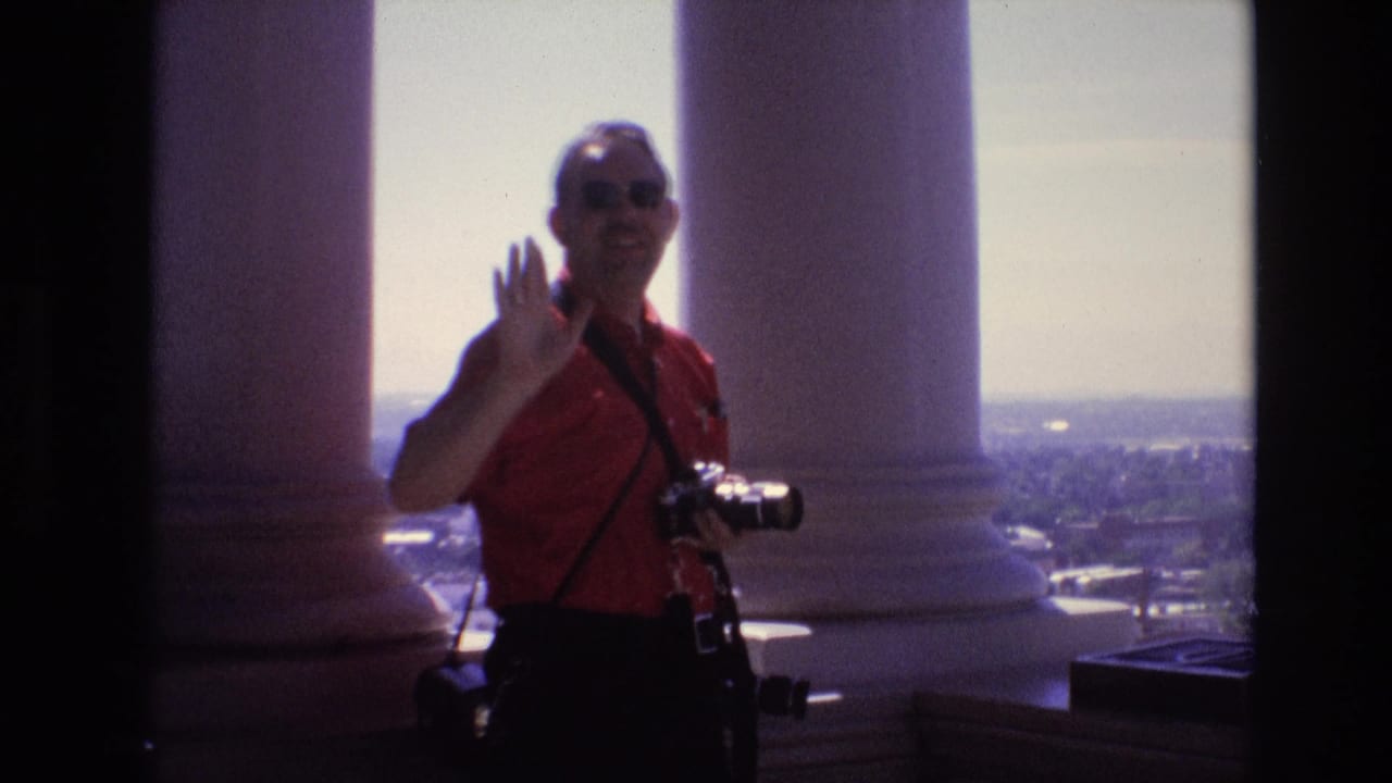 In Denver, Colorado in 1974, an adult man in sunglasses and a red shirt waves toward the filmmaker while holding a still camera between large white columns above the city.