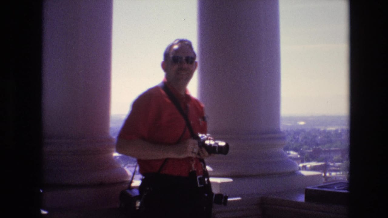 An adult man in a red shirt and sunglasses stands on a high Denver observation deck in 1974, holding a camera with white columns and the city skyline behind him.
