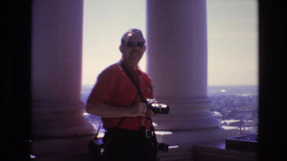 An adult man in a red shirt and sunglasses stands on a high Denver observation deck in 1974, holding a camera with white columns and the city skyline behind him.