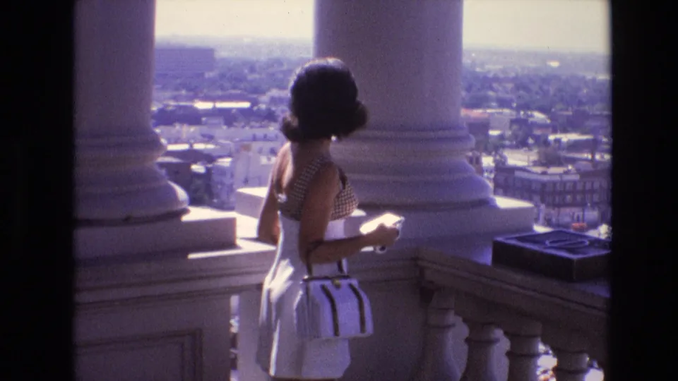 An adult woman in a checkered top, white skirt, and white purse stands near the balustrade of a Denver observation deck in 1974, looking toward the city view.