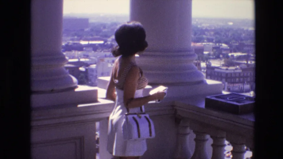 An adult woman with a white purse stands on a high observation deck in Denver in 1974, framed by white columns and a hazy city skyline in the background.