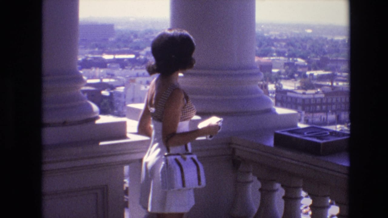 A woman in a white and patterned 1970s outfit stands near white columns and a stone balustrade, looking out over Denver from an observation deck.