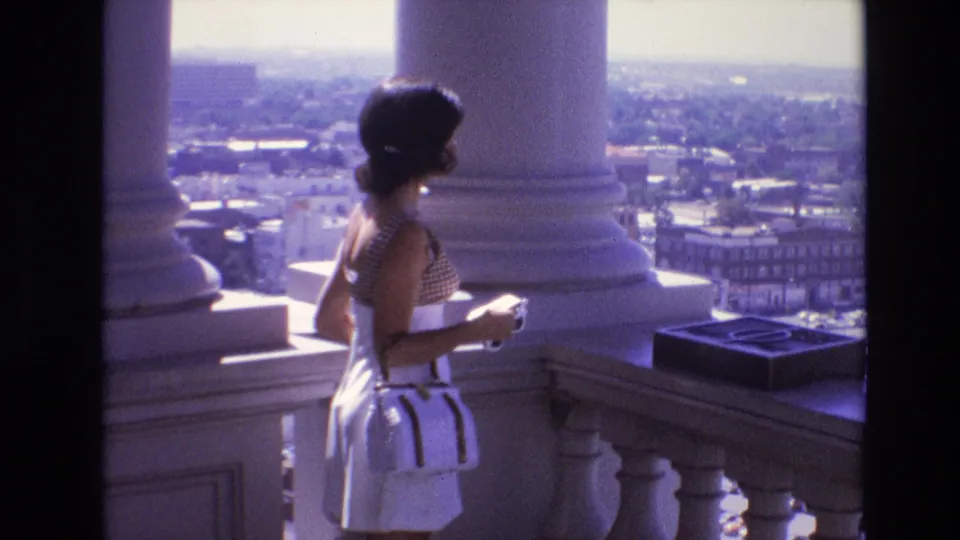 The woman holds a small camera while standing between large white columns above the Denver cityscape in the supplied 1974 home-movie footage.