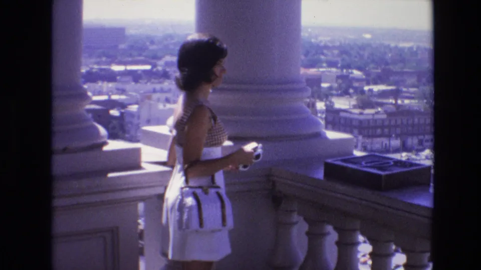A profile view shows the woman on the columned observation deck, with a stone railing, bright sky, and Denver urban buildings visible beyond her.