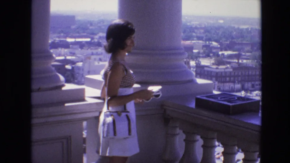 The woman remains at the stone balustrade with a camera in hand, framed by white columns and a broad Denver city view in 1974 home-movie footage.