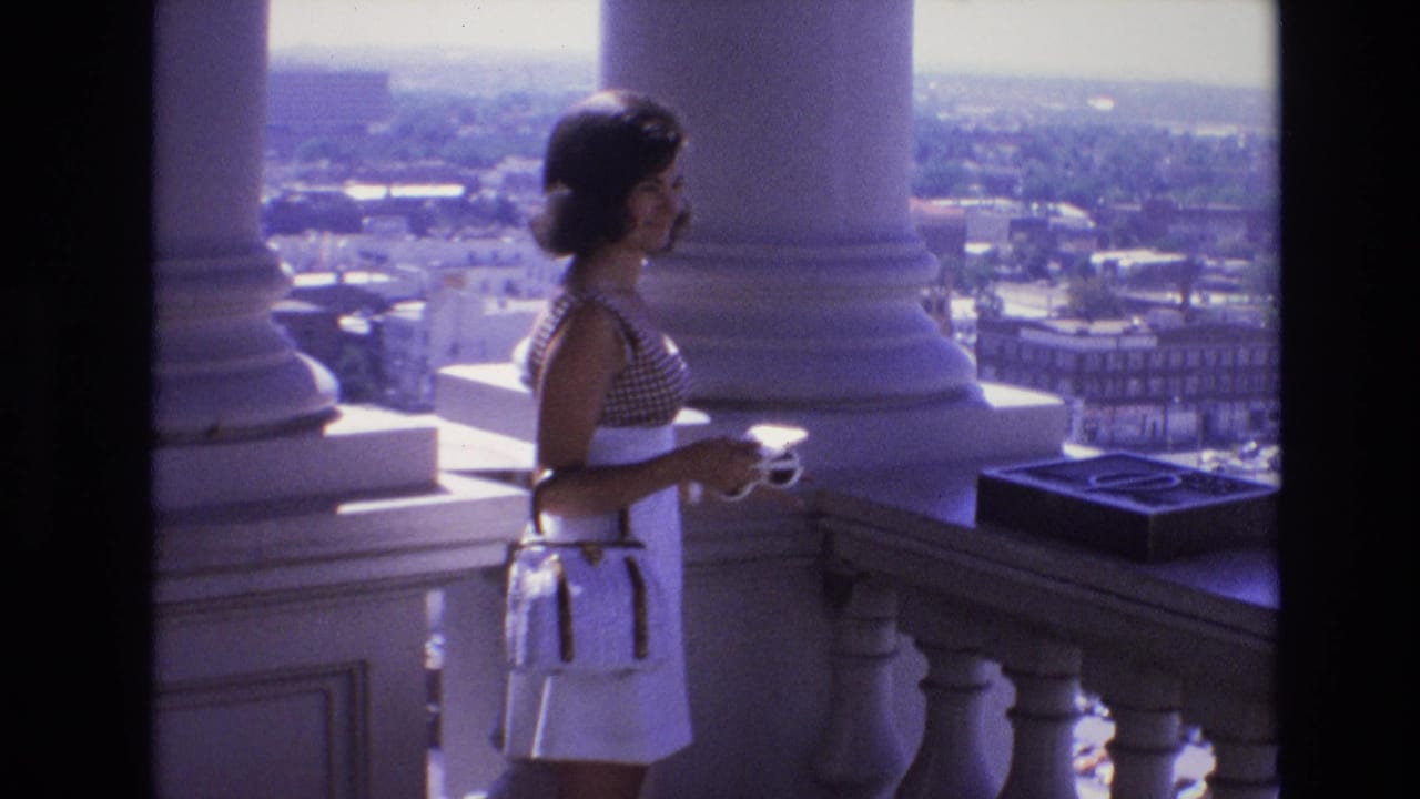 Young woman standing on a balcony beside large columns with a city view behind her.
