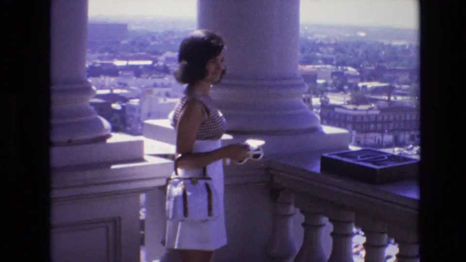 In supported 1974 Denver home-movie footage, a young woman in a checkered sleeveless top and white skirt stands on a high balcony beside classical columns, holding a small cup and handbag with the city visible beyond.