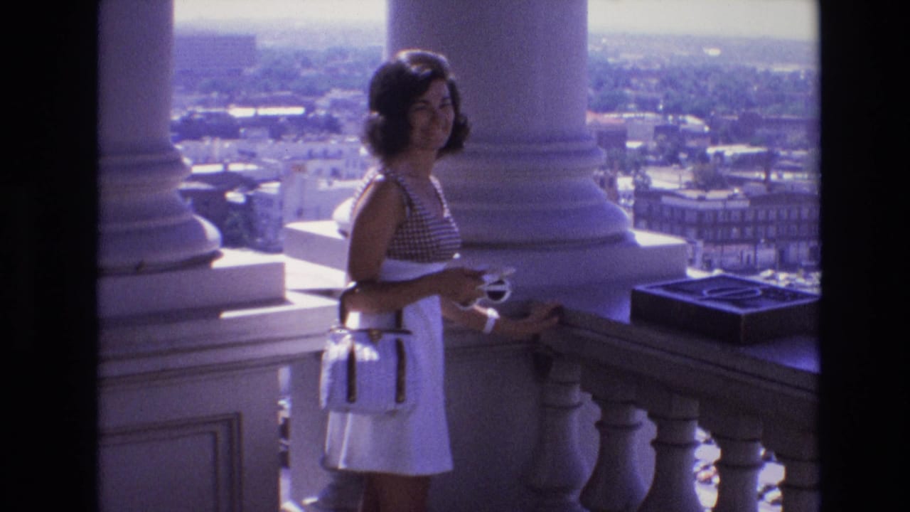 Woman on balcony turns toward the camera with columns and city buildings in the background.