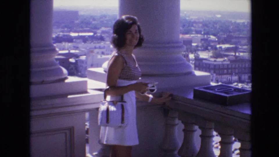 The young woman turns toward the camera on a columned balcony in Denver, holding a small cup near a stone railing as hazy city buildings stretch into the distance.