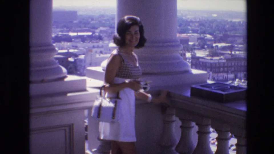 A smiling young woman in 1970s clothing faces the camera on a Denver balcony, framed by pale columns, a stone balustrade, and a soft distant urban view.