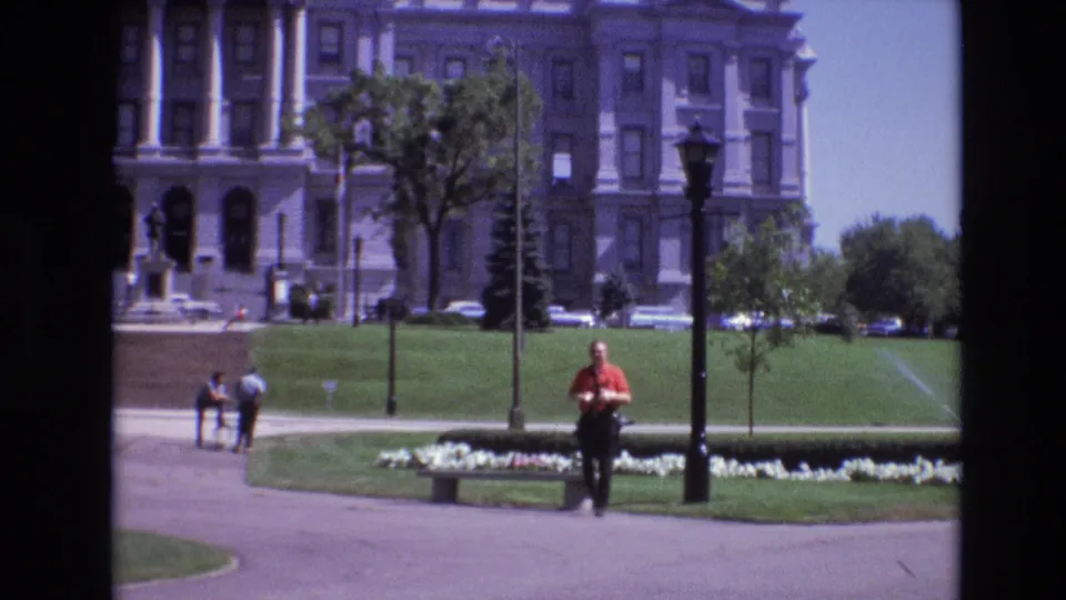 The scene shifts to a Denver public park view with green lawns, pedestrians, benches, lampposts, trees, flower beds, and a large historic-looking civic building in the background.