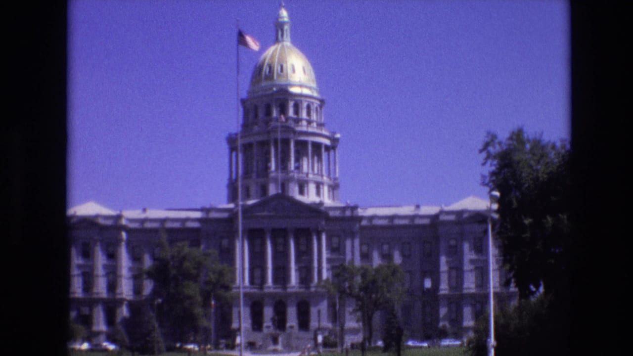 1974 8mm frame of the Colorado State Capitol in Denver, with the golden dome, columned entrance, American flag, trees, and lawn visible under a blue sky.
