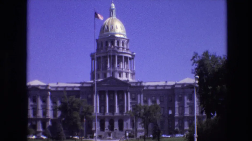 1974 8mm frame of the Colorado State Capitol in Denver, with the golden dome, columned entrance, American flag, trees, and lawn visible under a blue sky.