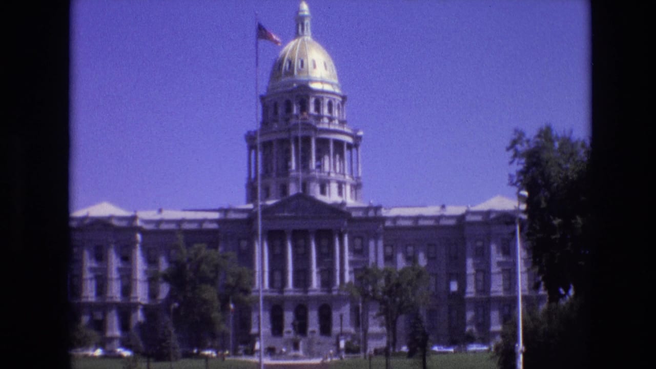 1974 Denver capitol view showing the golden dome, front columns, American flag on a tall pole, green lawn, and trees in soft 8mm color.