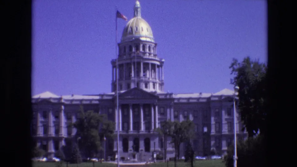 1974 Denver capitol view showing the golden dome, front columns, American flag on a tall pole, green lawn, and trees in soft 8mm color.