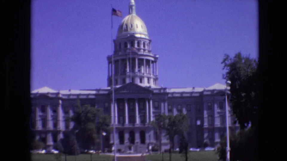 1974 home-movie frame of the Colorado State Capitol exterior, with the gold dome, broad stone facade, flagpole, trees, and foreground lawn visible.
