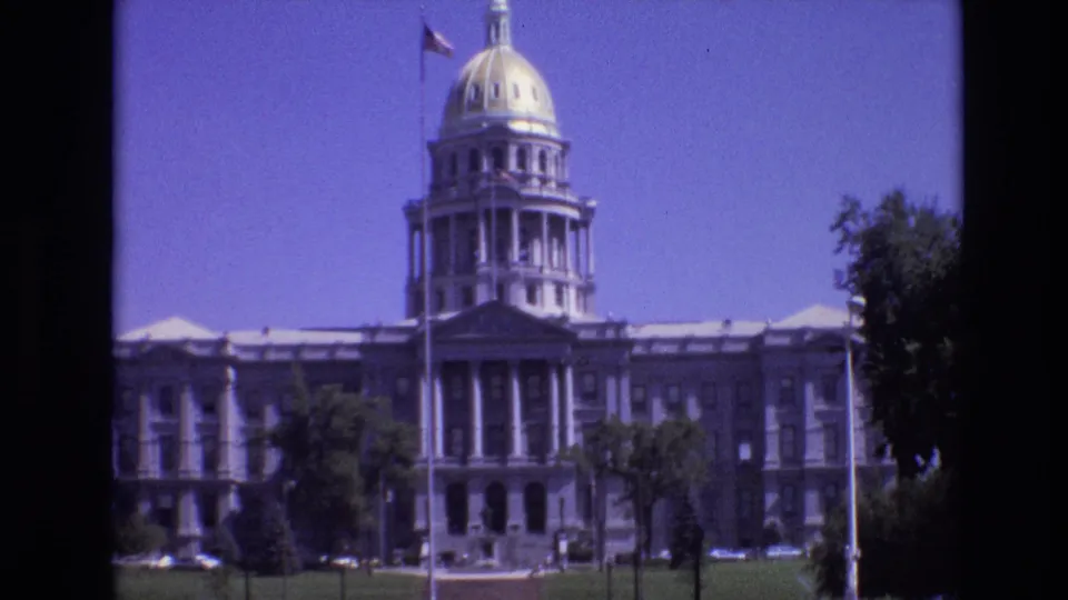 1974 8mm frame from Denver showing the Colorado State Capitol's dome and columned entrance, with an American flag, lawn, and trees in the foreground.