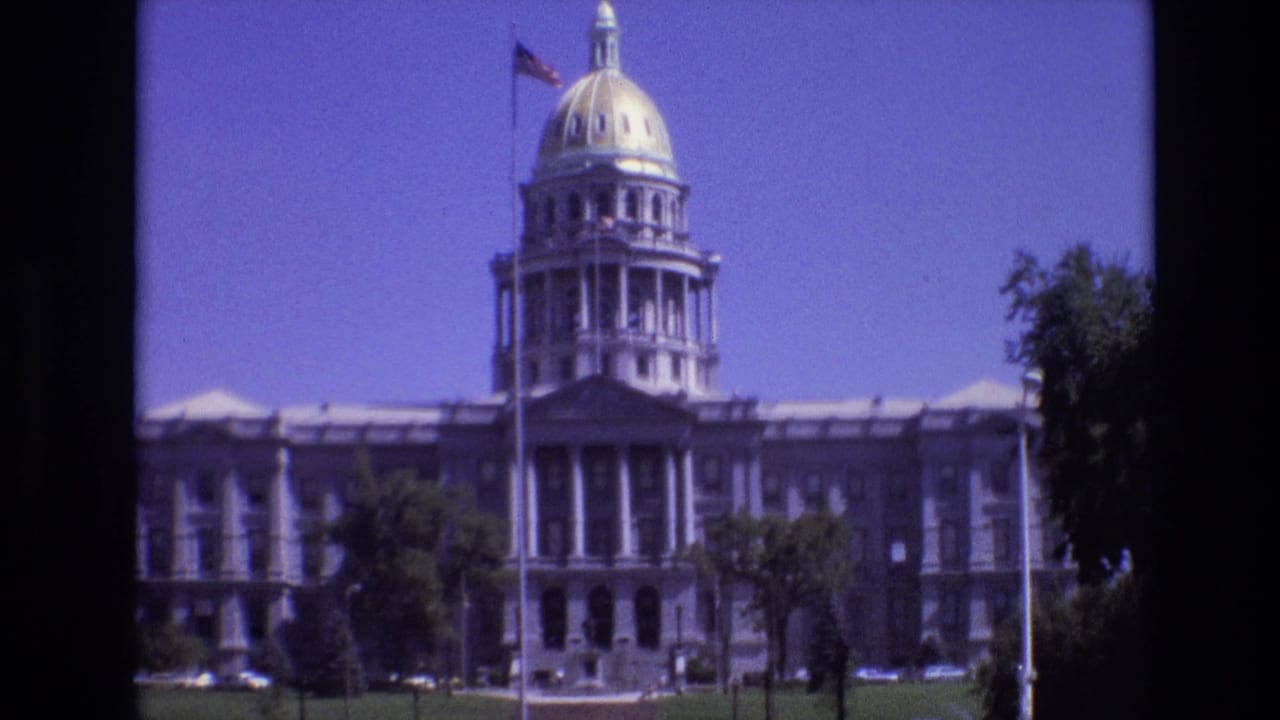 1974 Denver home-movie frame showing the Colorado State Capitol exterior with its gilded dome, American flag, front lawn, and surrounding trees.