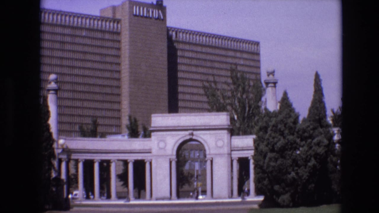 1974 Denver frame showing a Hilton hotel tower with visible rooftop branding behind a pale colonnaded entrance and landscaped trees.